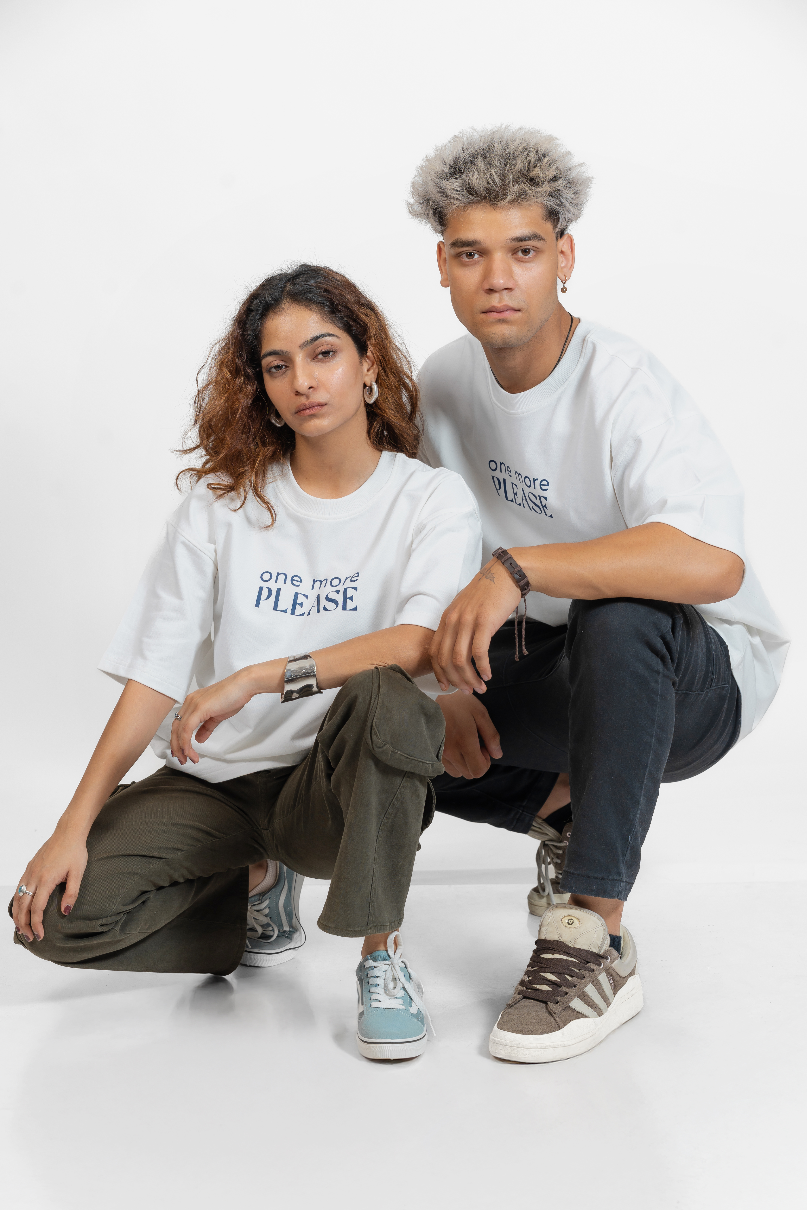 Two people wearing white t-shirts with text that says 'One More Please' in blue, sitting on a white background