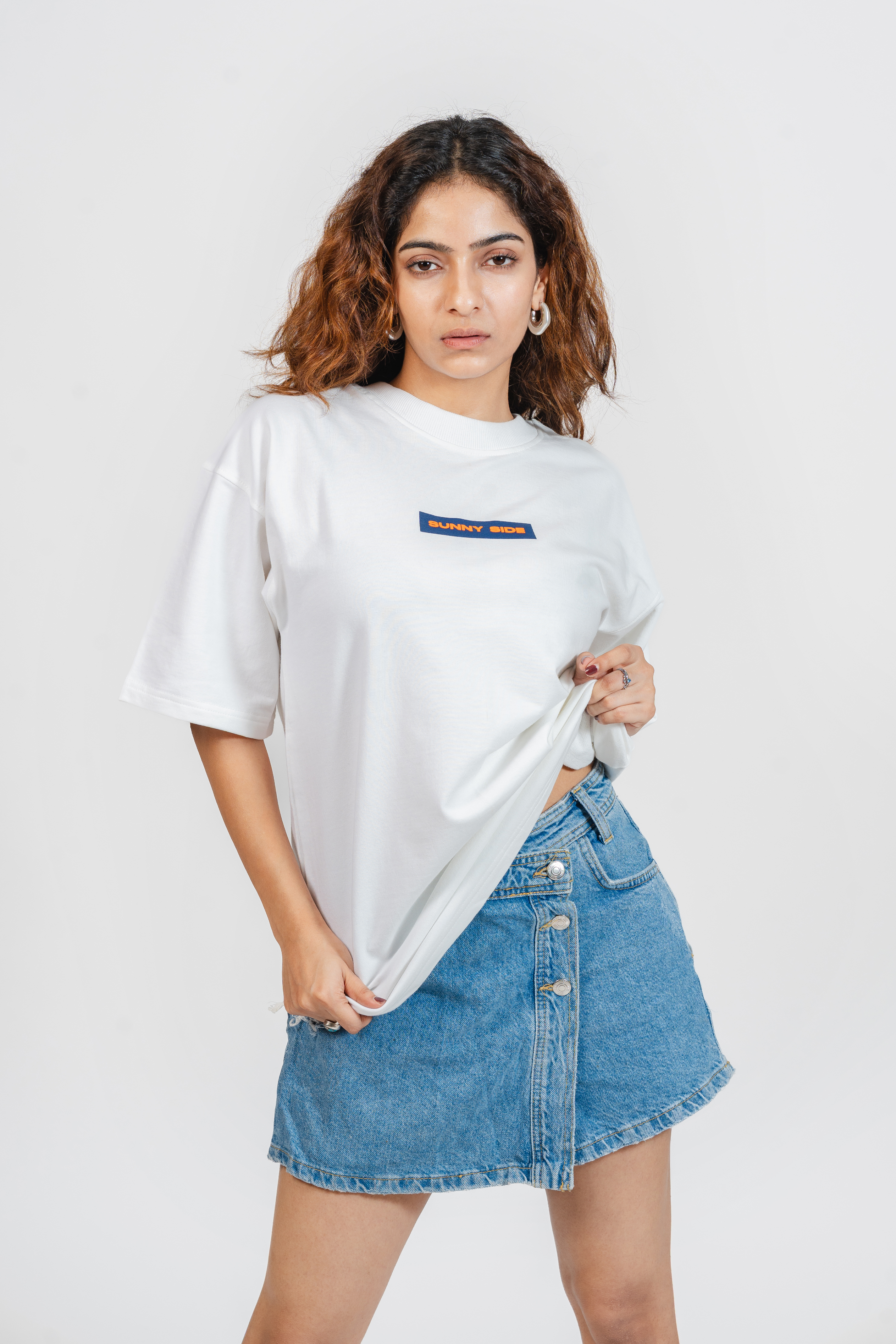 Woman wearing a white t-shirt with a small design on the front which says 'Sunny Side' and a denim skirt on a plain background