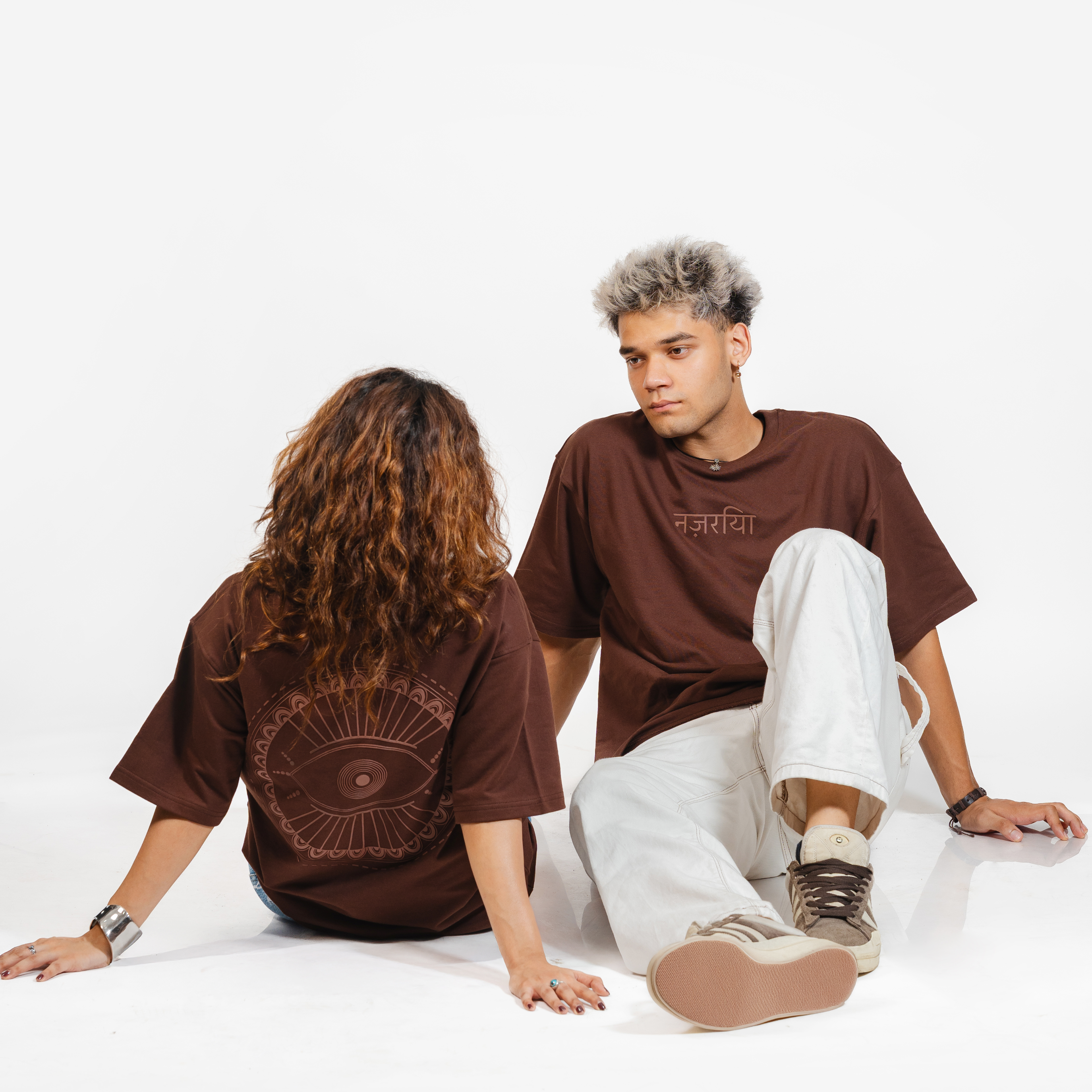 Man and woman wearing matching brown graphic t-shirts with Hindi text and eye design, seated on white background