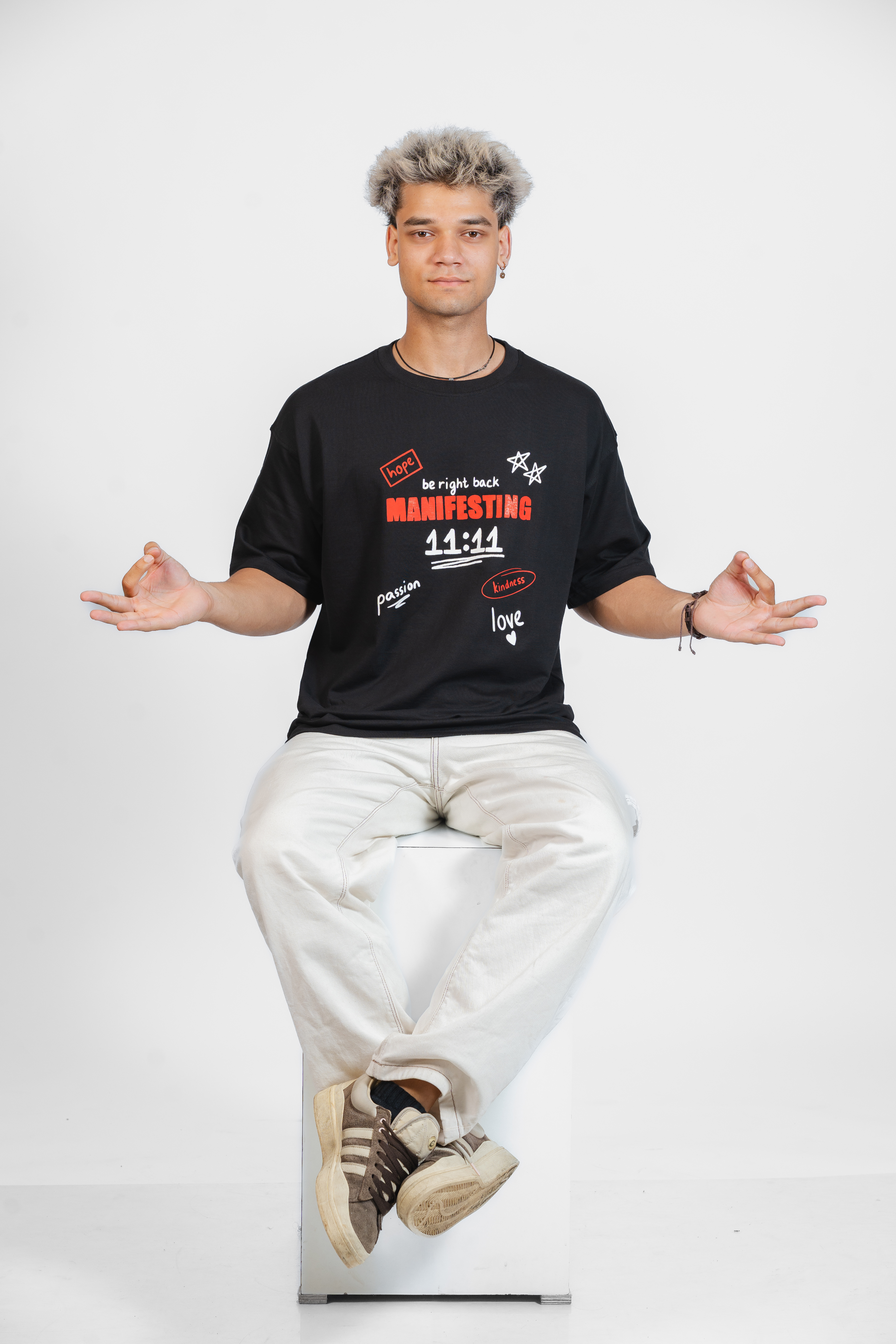 Man wearing a black t-shirt with text and graphics related to Manifesting, sitting on a white stool against a white background