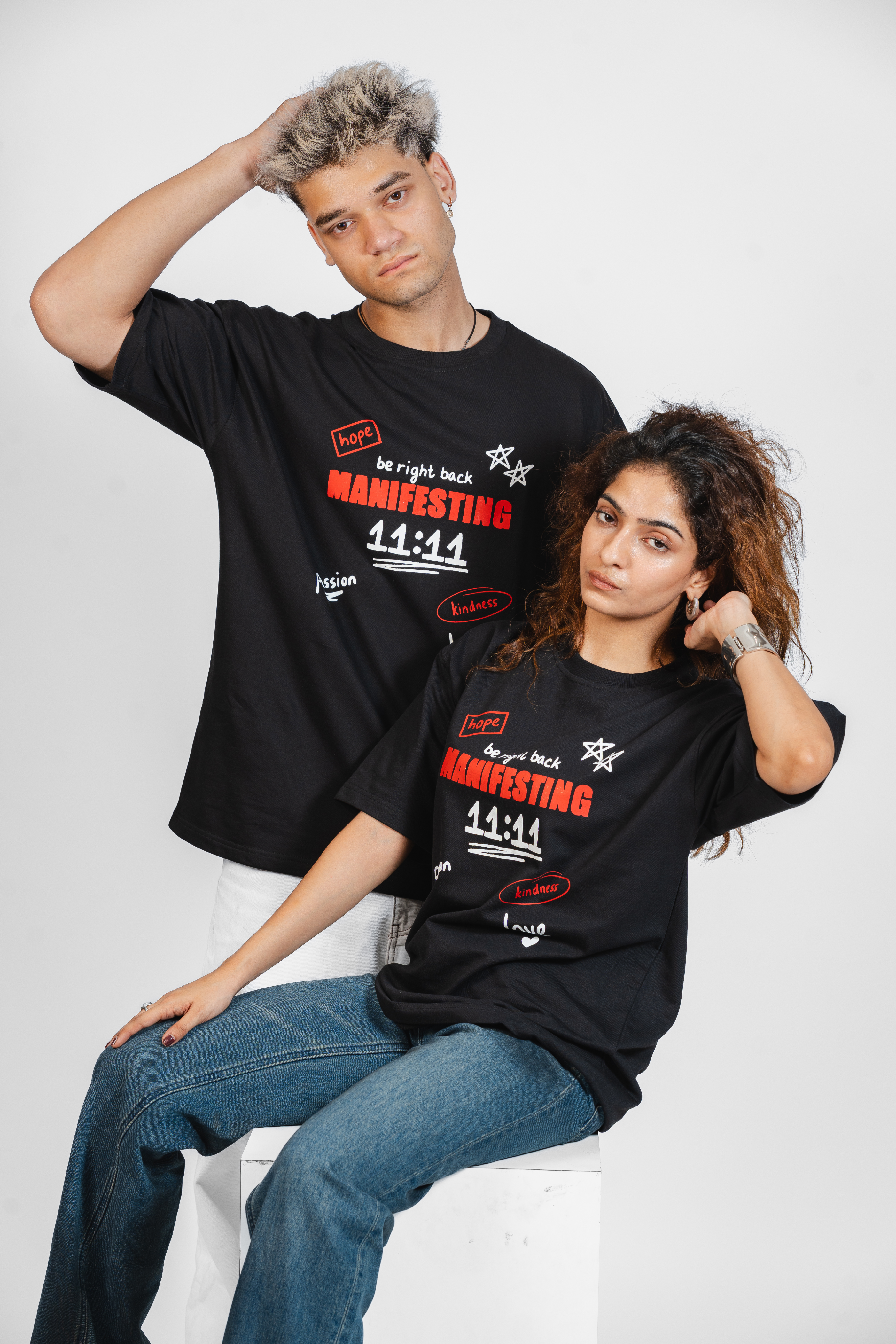 Two people wearing black t-shirts with red and white text related to 'Manifesting' and '11:11' on a white background