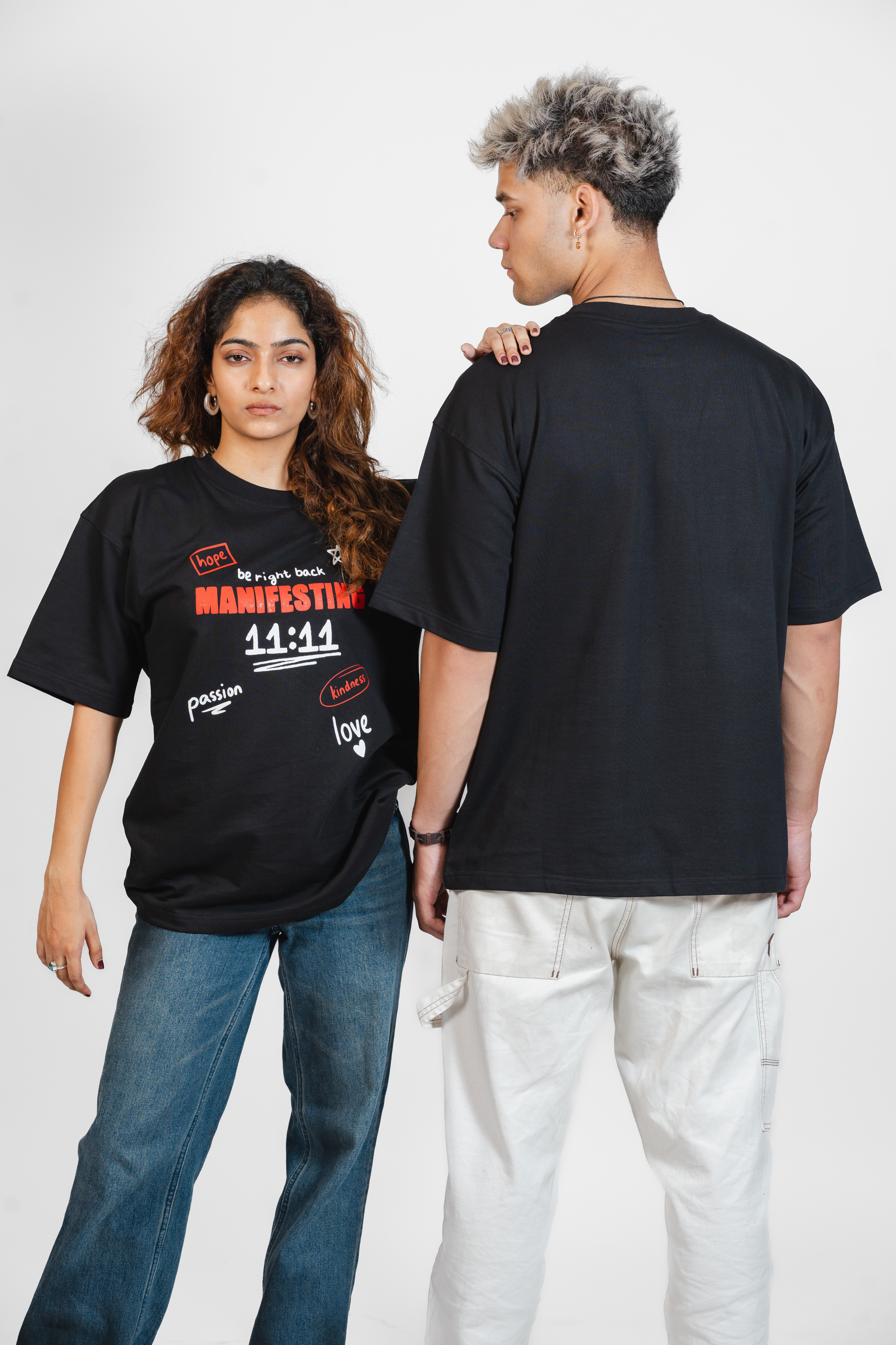 Two people wearing black t-shirts with text related to 'manifesting' and graphics on a white background