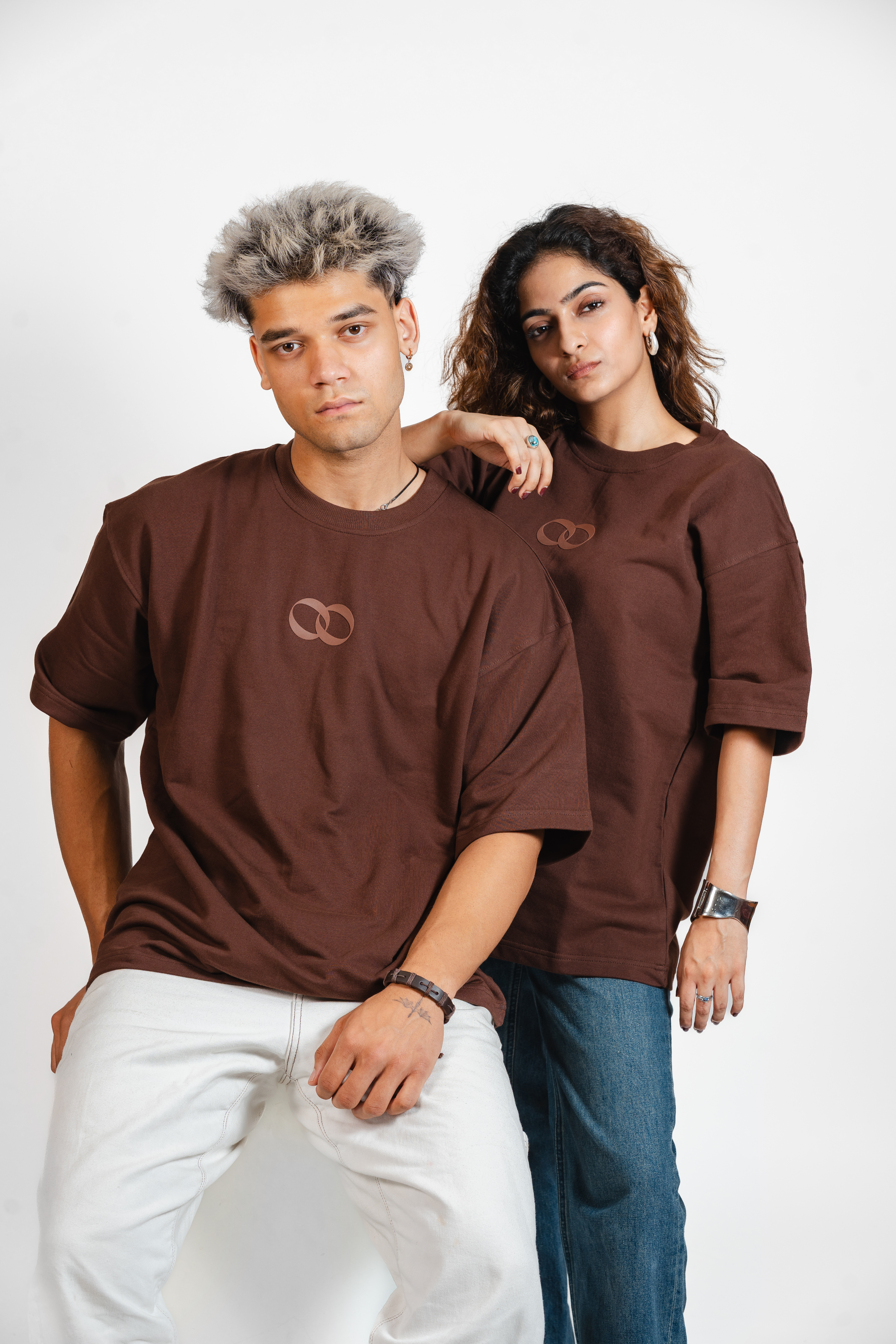 Two people wearing brown t-shirts with the one more please logo on a white backdrop