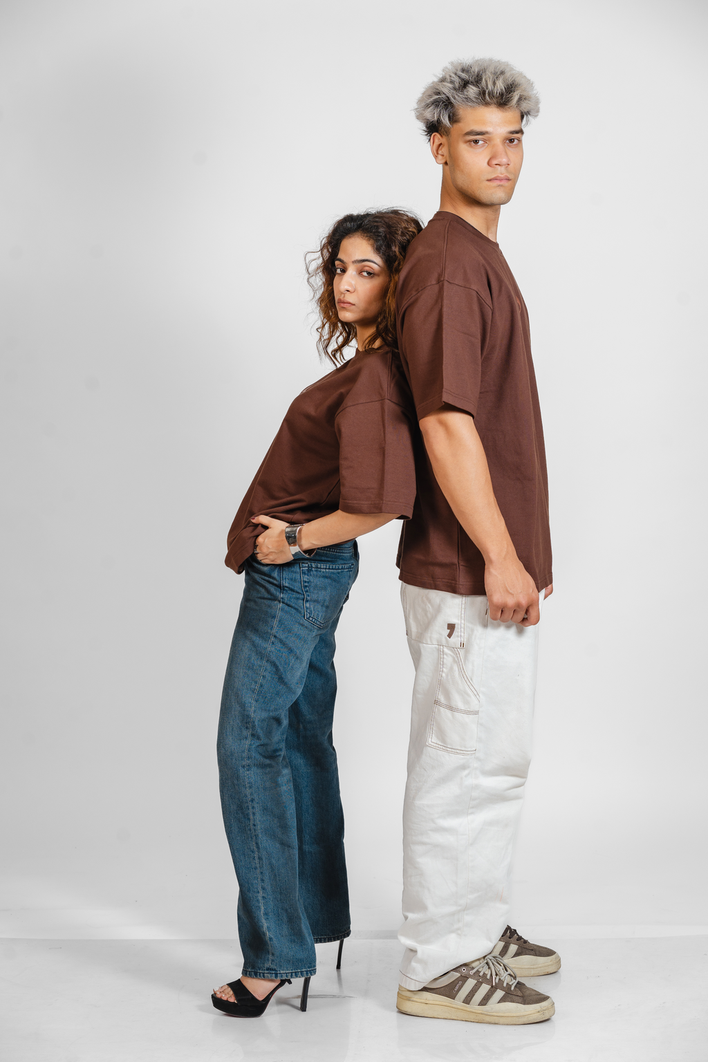 Two people wearing brown shirts and jeans standing back-to-back on a white background
