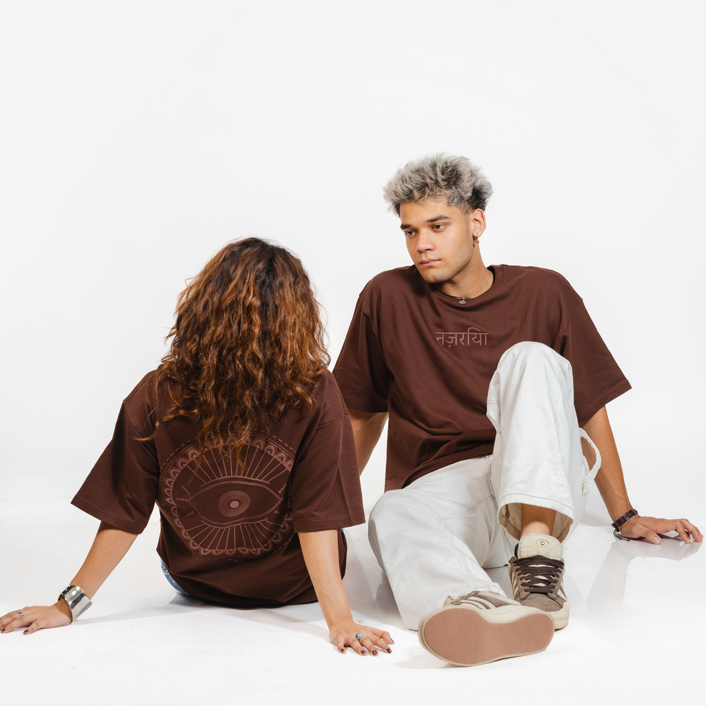 Man and woman wearing matching brown graphic t-shirts with Hindi text and eye design, seated on white background